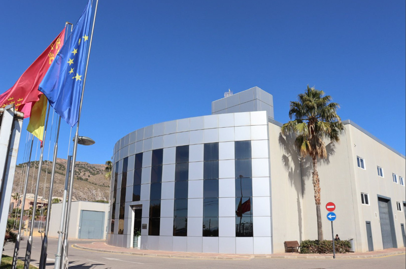 Edificio gris con cristaleras y delante la bandera de Europa, la de España, y la de Murcia.